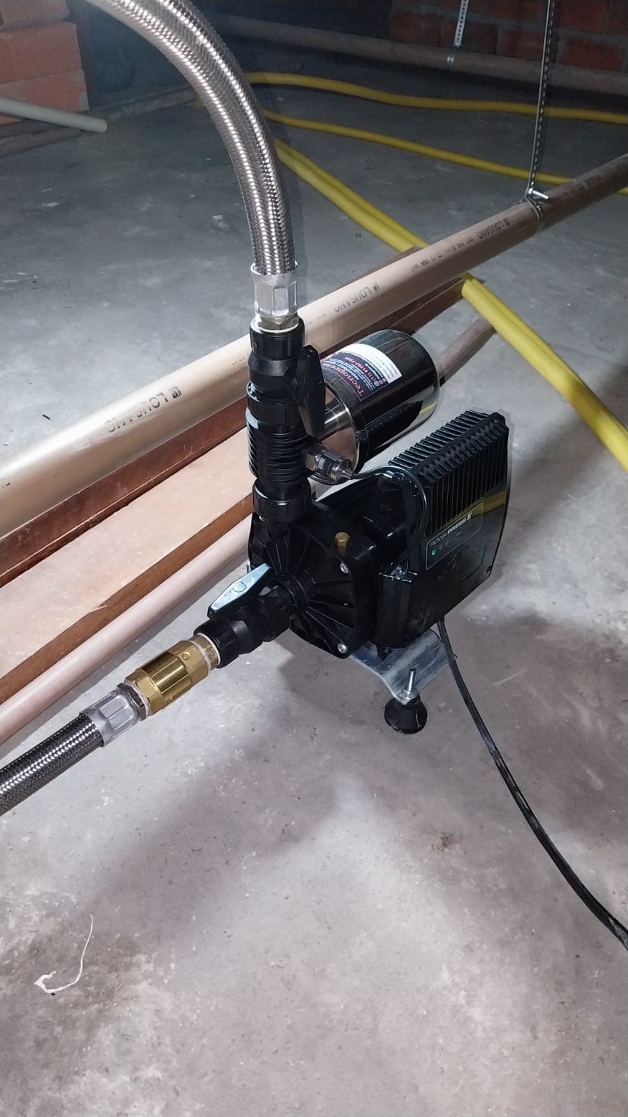 Pressurizador preto apoiado em uma base e conectado por mangueiras metálicas flexíveis.